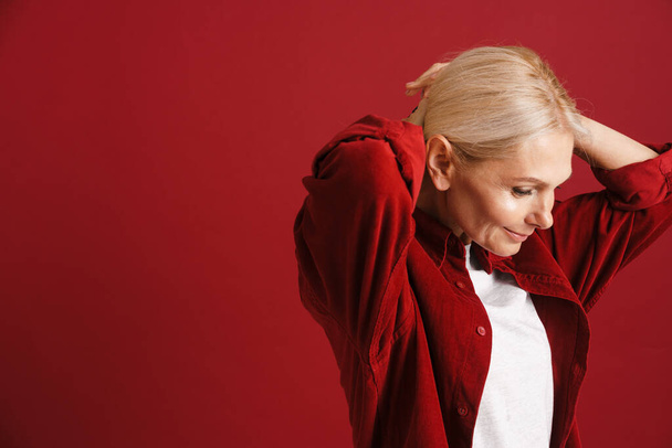 Mature blonde woman wearing shirt smiling and doing hairstyle isolated over red background - Φωτογραφία, εικόνα