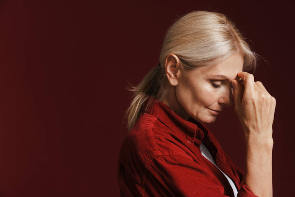 Mature blonde woman wearing shirt propping up her head isolated over red background - Fotó, kép