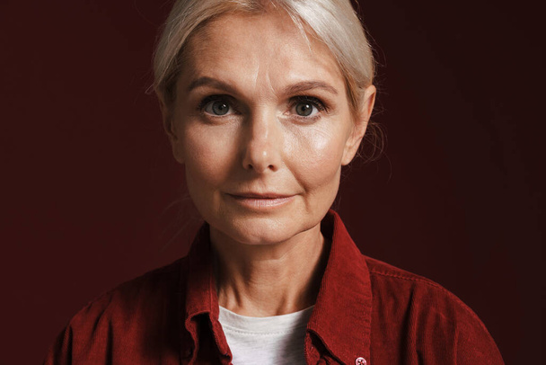Mature blonde woman wearing shirt posing and looking at camera isolated over red background - Foto, immagini