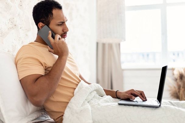 Arabic Guy Talking On Phone Using Laptop Working In Bedroom - Foto, afbeelding