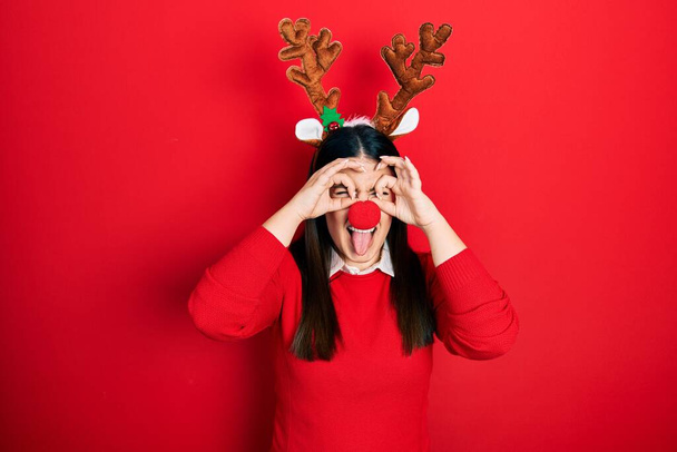 Mujer hispana joven con sombrero de Navidad de ciervo y nariz roja haciendo buen gesto como prismáticos sacando la lengua, ojos mirando a través de los dedos. expresión loca.  - Foto, Imagen