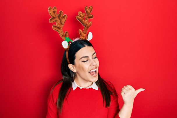 Jeune femme hispanique portant de jolies cornes de rennes de Noël souriant avec un visage heureux regardant et pointant vers le côté avec le pouce vers le haut.  - Photo, image