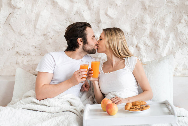 Loving young couple having breakfast in bed together, kissing, spending honeymoon at hotel - Foto, imagen