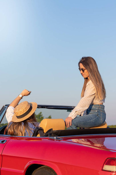 Two young women enjoying time together on a red classic convertible car on summer vacation time - Photo, Image