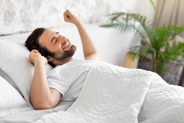 Zijaanzicht van lachende jongeman die zich uitstrekt in bed - Foto, afbeelding