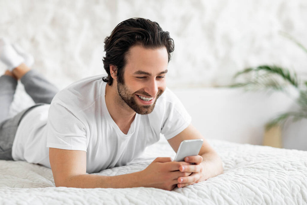 Iloinen kaveri juttelee ystävien kanssa älypuhelimella, makaa sängyssä - Valokuva, kuva
