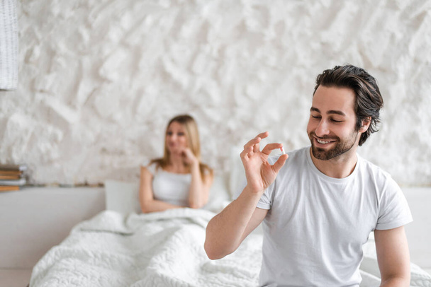Νεαρός άνδρας κρατώντας χάπι, επίλυση προβλημάτων σεξουαλικής δυσλειτουργίας, ευτυχισμένη γυναίκα κάθεται στο κρεβάτι και περιμένει τον άντρα της - Φωτογραφία, εικόνα
