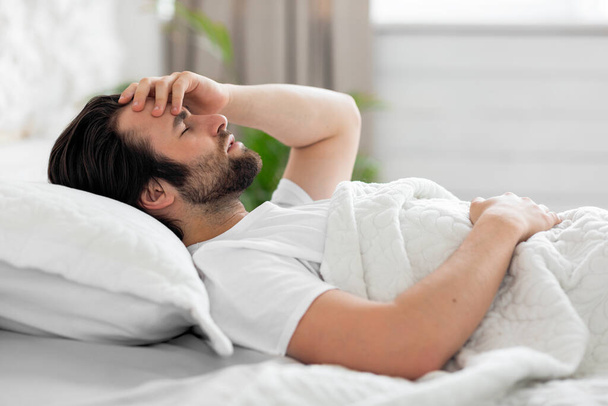 Upset young man waking up with headache, bedroom interior - Fotó, kép