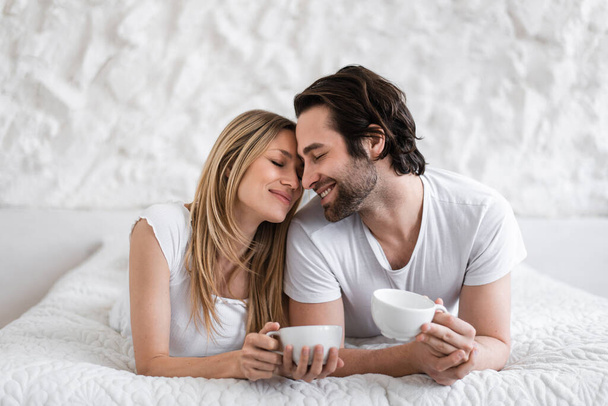 Romantic young couple lying on bed with cups of coffee, cuddling, spending time with each other at home - 写真・画像