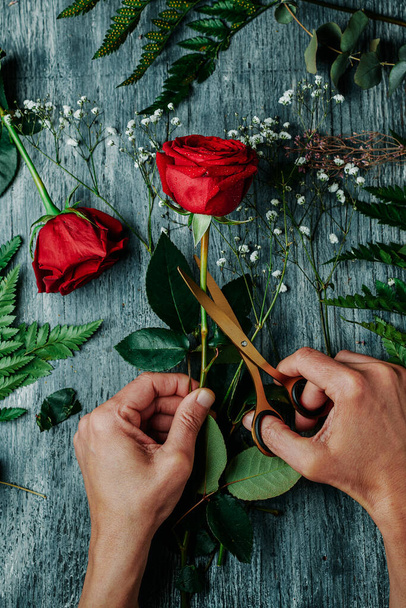 hoge hoek uitzicht van een man met behulp van een gouden schaar om een bloemstuk te maken, met een aantal rode rozen, op een grijs rustiek houten werkblad - Foto, afbeelding