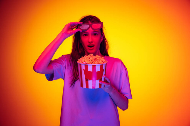 One excited young girl, student wearing 3d glasses and holding bucket of popcorn isolated on orange background in neon light, filter. Concept of emotions, youth, beauty - Fotografie, Obrázek