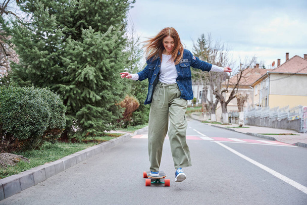 Lachende junge Skaterin auf der Straße in Bewegung mit einem Skateboard - Frontansicht einer modischen Skateboarderin in ihren Dreißigern - Foto, Bild