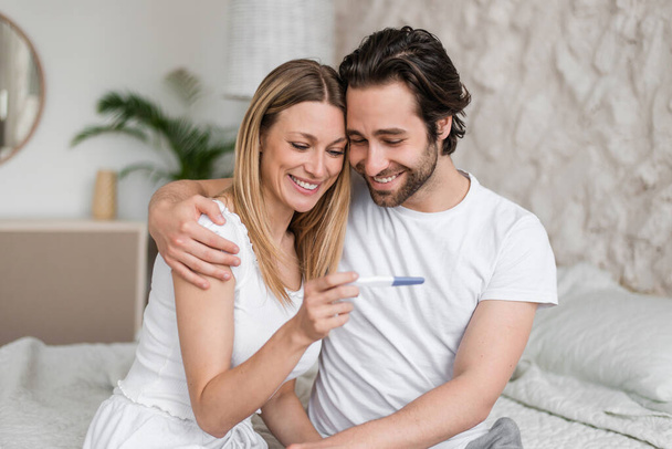 Cheerful millennial couple looking at positive pregnancy test, sitting on bed, hugging and enjoying great news - 写真・画像