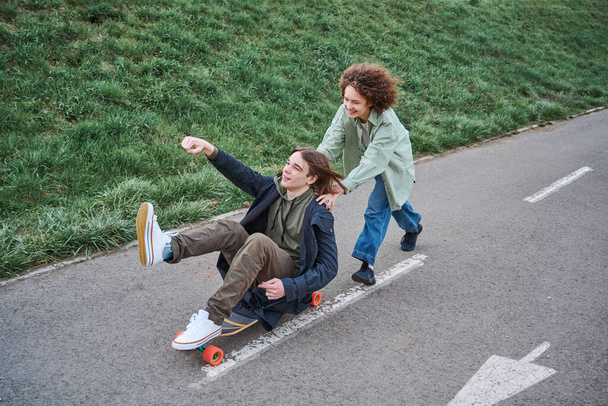 Glückliches Teenager-Mädchen schiebt lachenden Freund auf Skateboard. Freunde haben Spaß auf dem Skateboard in der Frühjahrs- oder Sommerpause. - Foto, Bild
