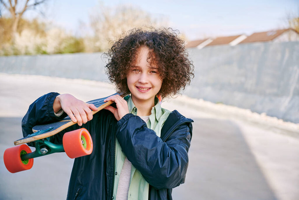 Sonriente adolescente patinadora femenina sosteniendo su monopatín. Chica con patinaje en el parque de skate mirando a la cámara al aire libre. - Foto, imagen