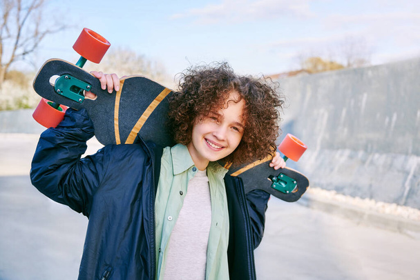 Uśmiechnięta nastolatka na deskorolce trzymająca deskorolkę. Dziewczyna z łyżwiarstwa w skate parku patrząc na kamery na zewnątrz. - Zdjęcie, obraz