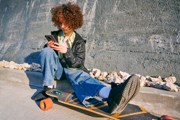 Znudzona nastolatka siedzi na deskorolce z telefonem komórkowym na świeżym powietrzu podczas letnich zajęć. Koncepcja psychologii młodzieżowej - Zdjęcie, obraz