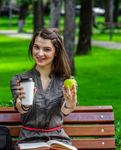 Young woman studying outside in a park holds a green apple and a cup of coffee looking to the camera.  - Zdjęcie, obraz