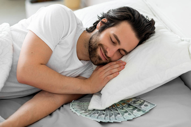 Smiling bearded guy sleeping with cash under his pillow - Foto, imagen