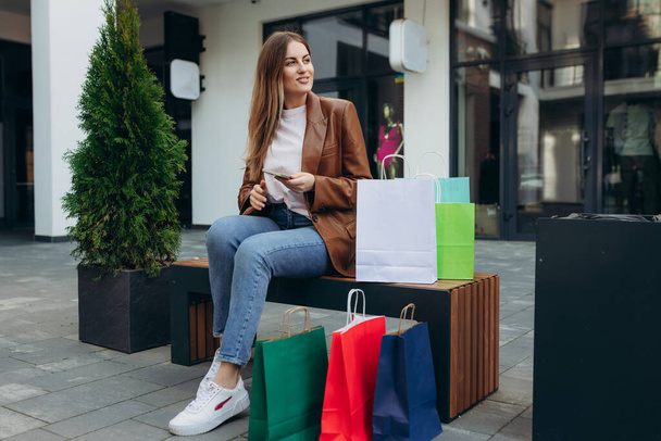Портрет елегантної молодої жінки, що тримає сумки для покупок і використовує смартфон під час виходу з торгового центру
 - Фото, зображення