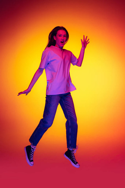 Full-length portrait of young cheerful girl, student with long hair jumping isolated on orange background in neon light, filter. Concept of emotions, youth, beauty - Фото, изображение