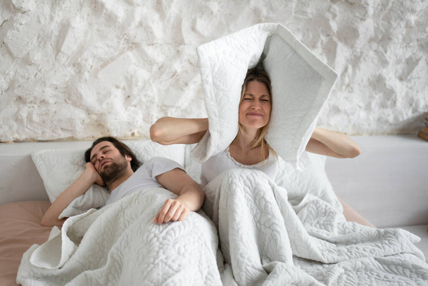 Frustrated young woman covering ears with pillow while her husband is snoring on bed at home, copy space - Foto, imagen
