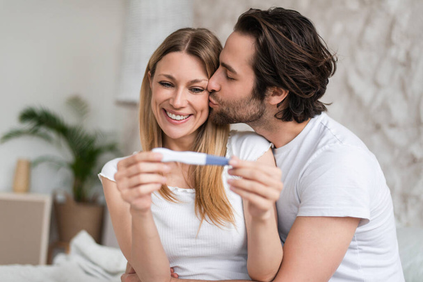 Loving young couple feeling happy about positive pregnancy test, kissing and hugging at home - Zdjęcie, obraz