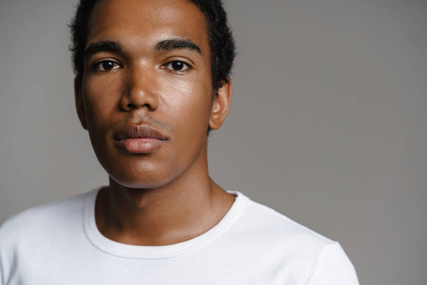 Young black man dressed white t-shirt posing and looking at camera isolated over grey wall - Foto, afbeelding