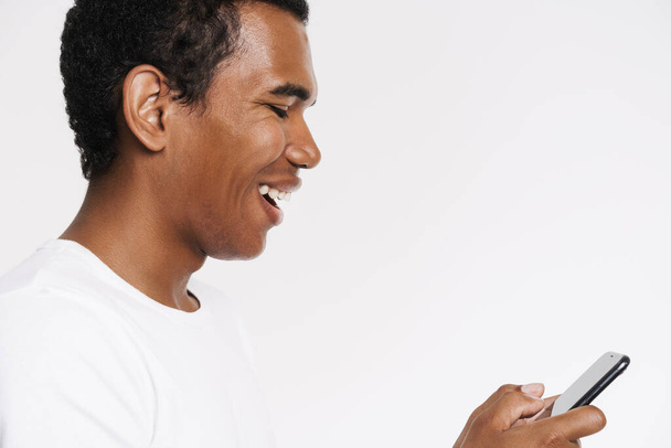 Young black man smiling while playing online game on cellphone isolated over white wall - Fotó, kép