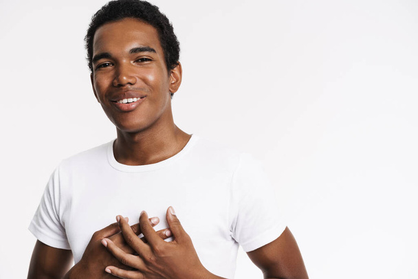 Young black man in t-shirt smiling while holding hands on his chest isolated over white background - Фото, зображення