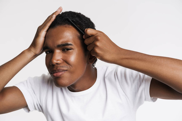 Young black man smiling at camera while combing his hair isolated over white wall - Фото, изображение