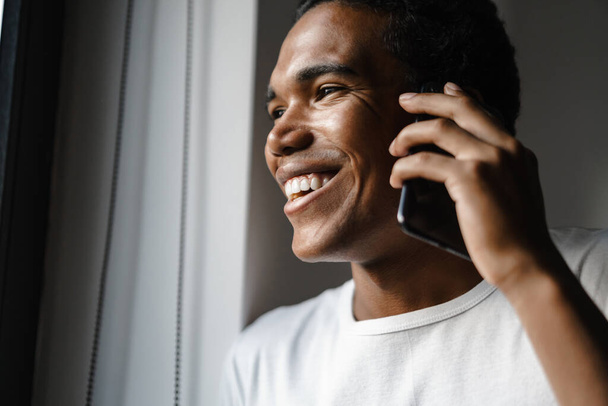 Young black man smiling and talking on cellphone while leaning on wall indoors - Photo, Image