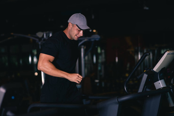 man wearing black sports shirt and cap running on treadmills and happy smiling in modern sport gym, cinematic dark tone  - Photo, Image