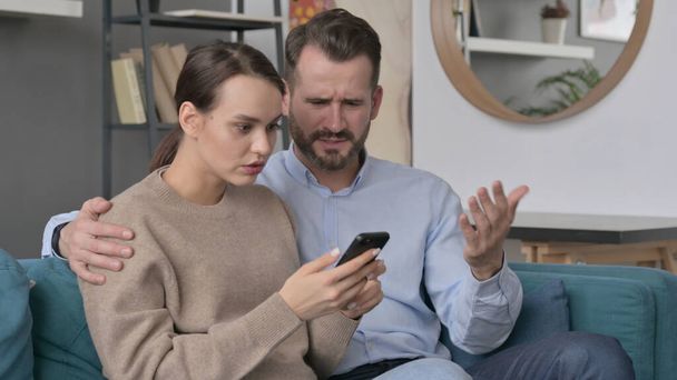 Portrait of Couple Reacting to Loss on Smartphone, Sitting on Sofa  - Fotografie, Obrázek