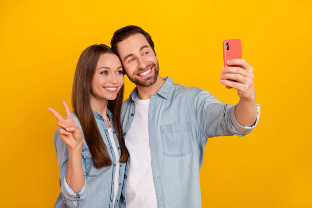 Photo of sweet young brown hairdo couple do selfie show v-sign wear jeans shirt isolated on yellow background - Zdjęcie, obraz
