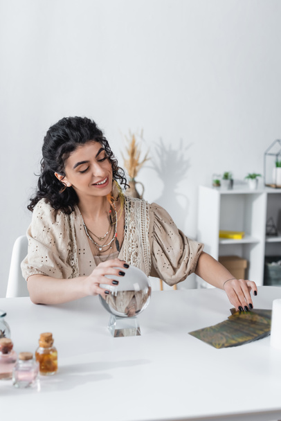 Smiling gypsy medium touching magic orb and tarot cards on table  - 写真・画像