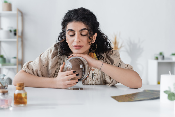 Young gypsy medium touching orb near blurred jars and tarot cards on table  - Photo, Image
