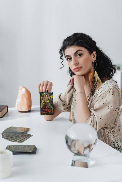 KYIV, UKRAINE - FEBRUARY 23, 2022: Gypsy fortune teller showing tarot card and looking at camera at home  - Photo, Image