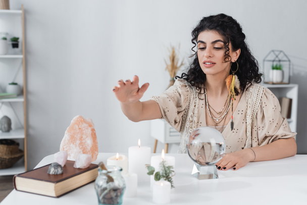 Gypsy fortune teller sitting near magic orb, candles and book at home  - Foto, Bild