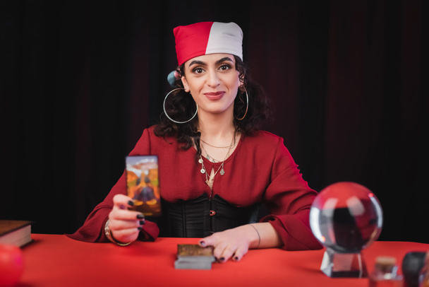 Smiling gypsy soothsayer holding tarot card near blurred orb isolated on black  - Foto, Imagem