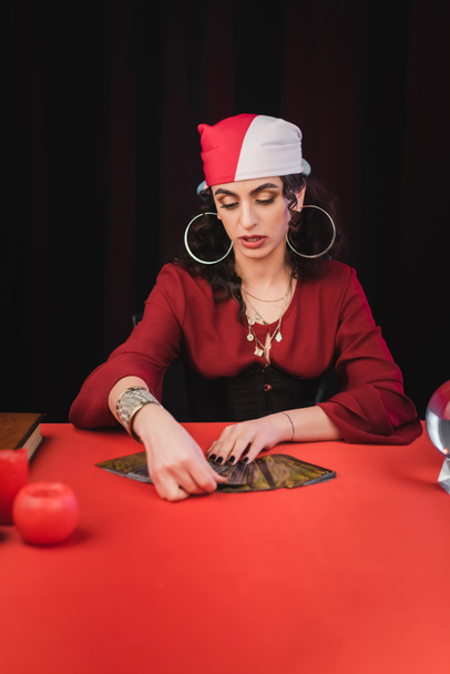 Gypsy soothsayer gospodarstwa karty tarota w pobliżu orb i książki na stole izolowane na czarno  - Zdjęcie, obraz