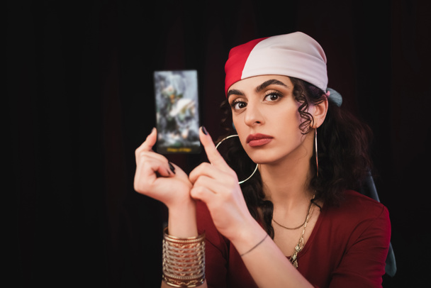 Gypsy fortune teller holding tarot card and looking at camera isolated on black  - Foto, Imagen