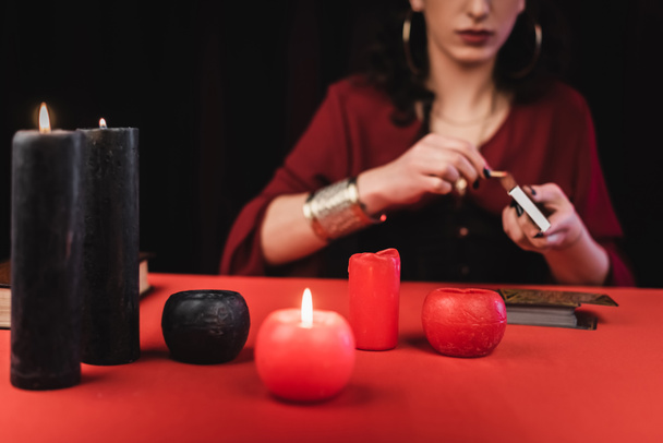 Cropped view of blurred fortune teller holding matches near candles and tarot cards isolated on black  - Fotografie, Obrázek