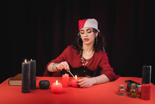 Gypsy soothsayer burning candles near book and witchcraft supplies on table isolated on black  - Fotoğraf, Görsel