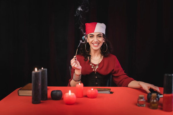 Cheerful gypsy soothsayer holding match near candles and blurred witchcraft supplies isolated on black  - Foto, Imagen