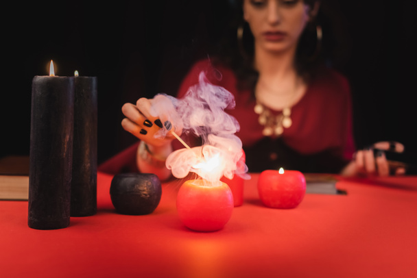 Cropped view of blurred soothsayer burning red candle on table isolated on black  - Fotografie, Obrázek