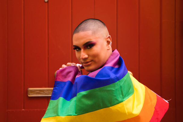 Non-binary person, of South American origin and young, the person is wearing make-up and carries the gay pride flag on his shoulders. Concept equality, homosexuality, gay, lesbian, gay pride. - Valokuva, kuva