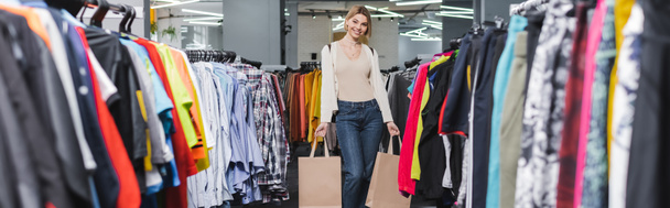 Smiling woman holding shopping bags near clothes in second hand, banner  - Foto, Bild