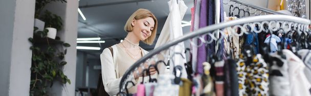 Positive woman touching scarf near rack in second hand, banner  - Photo, Image