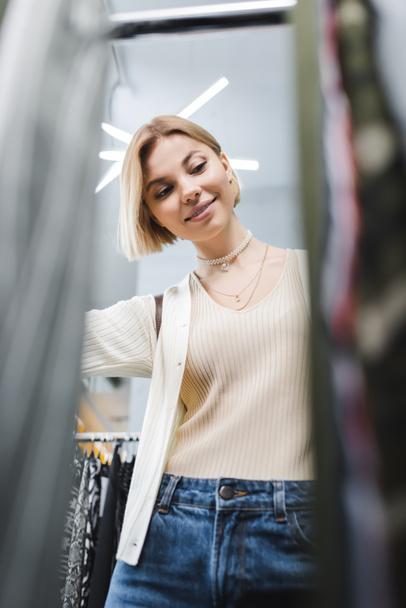Low angle view of positive woman looking at blurred clothes in second hand  - Foto, afbeelding
